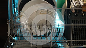 Close-up cropped shot of unrecognizable housewife loading backlit dishwasher machine with glass cup at evening. Concept