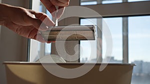 Close-up cropped shot of unrecognizable man cleaning filter of modern robot vacuum cleaner using brush by window on