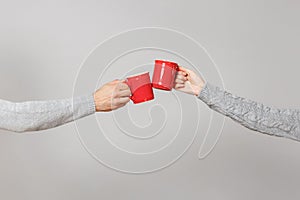 Close up cropped of woman, man two hands horizontal holding red cups of tea, clinking isolated on grey wall background