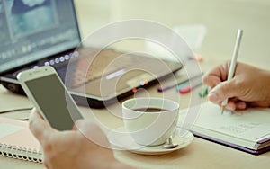 Close up cup of coffee and laptop computer with hand of business man using smart phone.