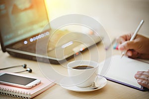 Close up cup of coffee and smart phone with hand of business man using laptop computer and write notebook on wooden desk office w