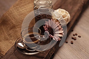 Close up of a cup of coffee on a wooden board, with a flower and a container of coffee beans.