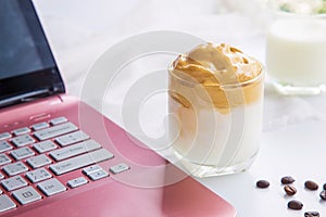 Close up a cup of Dalgona coffee with coffee beans next to a pink laptop on a white table with a cup of milk blurred background