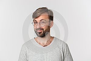 Close-up of curious bearded man in gray t-shirt and glasses, smiling intrigued at camera, standing over white background