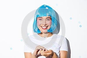 Close-up of cute asian girl in blue wig celebrating halloween, throwing confetti and smiling, standing over white