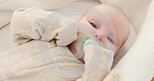 Close-up of a cute baby with a pacifier in his mouth, lying on a white blanket, sobbing.