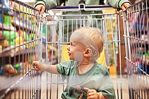 Close-up of cute baby in shopping cart being driven by her mom in supermarket. Mother and little blonde daughter in