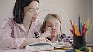 Close-up of cute caucasian girl reading book with mom at home. Happy schoolgirl doing homework with parent. Mother