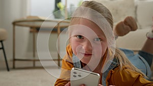 Close-up cute caucasian little kid girl lying on floor in living room using mobile phone application happy child playing