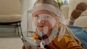 Close-up cute caucasian little kid girl lying on floor in living room using mobile phone application happy child playing