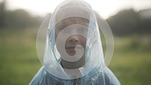 Close-up of cute charming boy in raincoat hood standing in sunrays on rainy summer day. Portrait of confident Caucasian