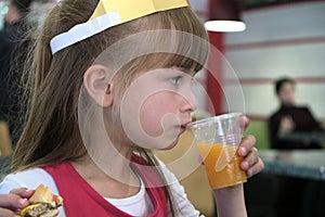 Close up of cute child girl drinking orange juice from plastic cap and eating fast food in a restaurant