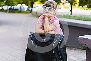 Close-up of a cute happy little traveler girl in sunglasses leaning on a black suitcase. Smiling pretty small girl with a suitcase