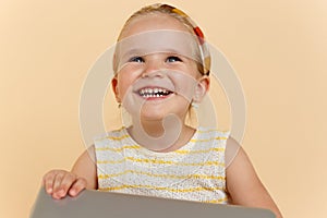 Close up of a cute little girl smiling and looking up, holding a laptope, over beige background. Horizontal view.