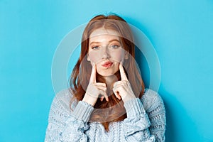 Close-up of cute redhead girl in sweater, pucker lips and pointing fingers at cheeks, poking dimples, standing over blue