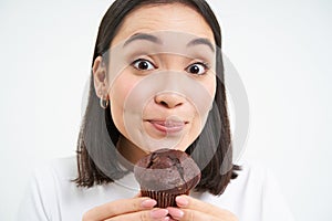 Close up of cute smiling asian woman, holding chocolate cupcake near mouth, having bite, enjoys eating pastry, white