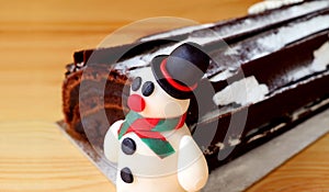 Close-up of a Cute Snowman Marzipan with Blurred Chocolate Yule Log Cake in Background Isolated on Wooden Table