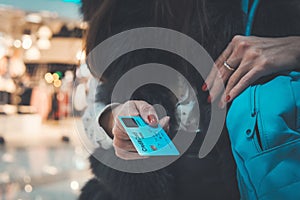 Close up of dark-haired girl in fur waistcoat holding credit card and bag at mall background. Stylish young woman paying