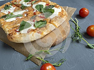 Close-up of delicious fresh crispy Roman pizza with cheese and herbs on a gray background. Fresh ripe tomatoes are nearby. Craft