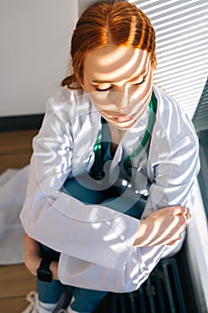 Close-up of depressed sad young female doctor in white coat sitting on floor hugging legs with hands near window.