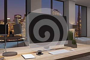 Close up of designer office desktop with empty black computer monitor, window with night city view, decorative items and supplies