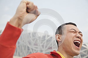 Close up of determined young man in athletic clothing with fist in the air, with modern building in the background in Beijing, Chi