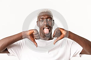 Close-up of disappointed african-american blond male showing thumbs-down and stick tongue from disappointment, dislike