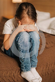 Close-up of disappointed little girl hugging knee, sobbing with head bowed and crying sitting alone on bed in bedroom.