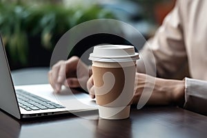 Close-up of a disposable coffee cup on a desk with a person typing on a laptop.