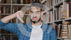 Close-up dissatisfied upset indian man student looking at camera standing in library showing thumb down demonstrating