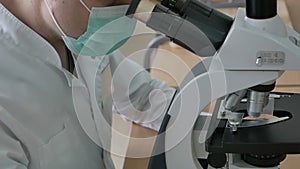 Close-up of a doctor looking through a microscope in a bandage in a doctor`s uniform. Taking biomaterial for analysis Covid -19. M