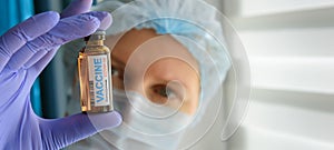 Close up doctor in medical mask and latex gloves holds in her hand vial with covid-19 vaccine. Nurse in white coat  looks at the