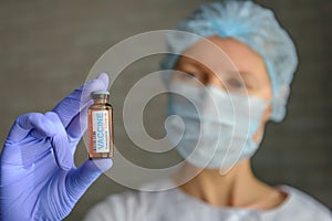 Close up doctor in medical mask and latex gloves holds in her hand vial with covid-19 vaccine. Nurse in white coat  looks at the