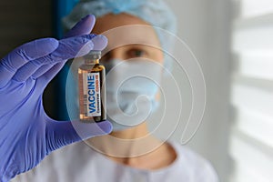 Close up doctor in medical mask and latex gloves holds in her hand vial with covid-19 vaccine. Nurse in white coat  looks at the