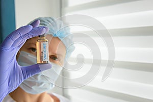 Close up doctor in medical mask and latex gloves holds in her hand vial with covid-19 vaccine. Nurse in white coat  looks at the