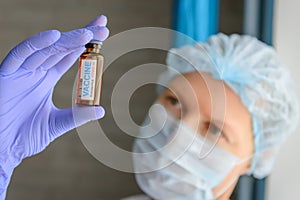 Close up doctor in medical mask and latex gloves holds in her hand vial with covid-19 vaccine. Nurse in white coat  looks at the