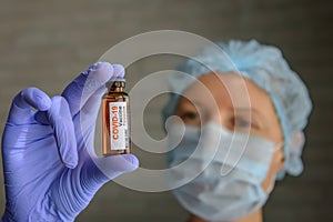 Close up doctor in medical mask and latex gloves holds in her hand vial with covid-19 vaccine. Nurse in white coat  looks at the