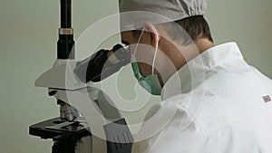 Close-up of a doctor in uniform in a mask in the laboratory examines a biomaterial under a microscope. Research ea viruses. Medica