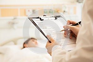 Close up of doctor writing on a medical chart with patient lying in a hospital bed in the background
