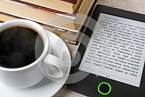 Close-up of e-book reader near cup of hot coffee and stack of ordinary paper books. Copy space on e-book display. E-reading for
