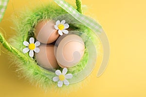 Close up. Easter card. Rustic eggs in a green basket decorated with bird`s fluff. Yellow background. Felt flowers. Copy space
