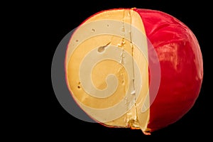 Close up. Edam cheese head in a cut. Yellow cheese on top covered with a red wax shell. Isolated on black background