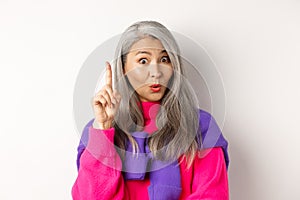 Close up of elderly asian woman in stylish hipster clothes, raising finger and saying idea, have suggestion, standing
