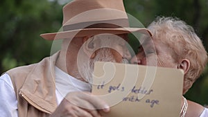 Close-up. An elderly husband and wife look at each other and kiss, hiding behind a sign with the inscription it gets