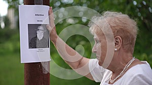 Close-up. An elderly woman posts a paper notice about a missing elderly man on a pole in a city park. The woman is upset