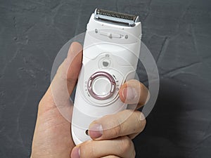 Close up Electric epilator in white color in a man`s hand on a gray textured background. Means for hair removal, beauty care,