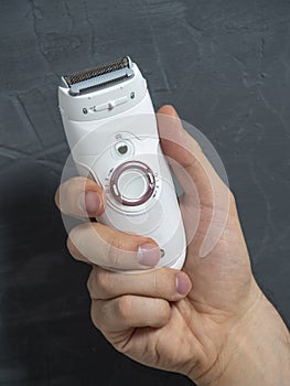 Close up Electric epilator in white color in a man`s hand on a gray textured background. Means for hair removal, beauty care,