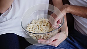 Close-up of elegant couple`s hands holding a big transparent bowl and eating popcorn at home. Couple wering blue jeans