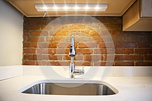 Close-up of a elegant, silver kitchen sink with faucet, white counter top, led light and red brick wall