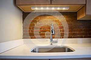 Close-up of a elegant, silver kitchen sink with faucet, white counter top, led light and red brick wall